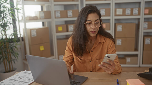 Woman holding smartphone and typing on laptop with bare hands among stacked parcel boxes on shelving in a building office; focused productivity. - Powered by Adobe