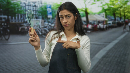 Woman holding whisk and pointing finger to whisk on a cobblestone street cafe; skepticism culinary humor.