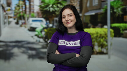 Brunette woman with a volunteer shirt standing with crossed arms outdoors on a sunny street, conveying community involvement and brightness in an urban environment.