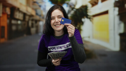 Woman in purple volunteer shirt on street contrasts emotions holding us dollars and credit card,...