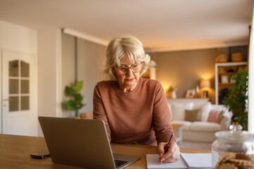 Elderly woman writing expenses in notepad and using laptop at desk while managing budget in living room