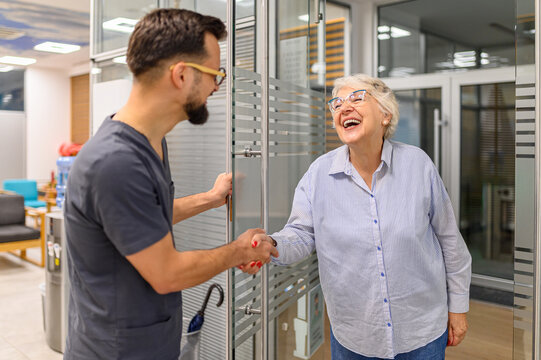 Male ophthalmology doctor greeting happy senior woman with handshake while opening glass door at clinic entrance