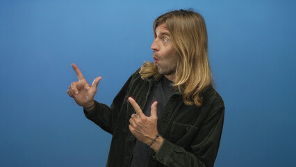 Blond man with long hair pointing both index fingers to right in blue studio with open mouth...