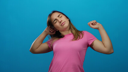 Fototapeta premium Woman stretching with closed eyes isolated on a bright blue background, expressing relaxation, wearing a pink shirt, blonde hair, young adult, peaceful, casual expression.