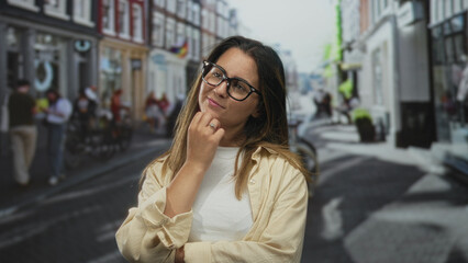 Fototapeta premium Woman touching chin with hand on a city street wearing glasses and casual shirt; contemplation quiet curiosity.