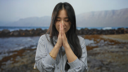 Young woman praying on a serene beach with a peaceful sea backdrop, captured in quiet meditation, symbolizing tranquility and contemplation amidst nature's beauty.
