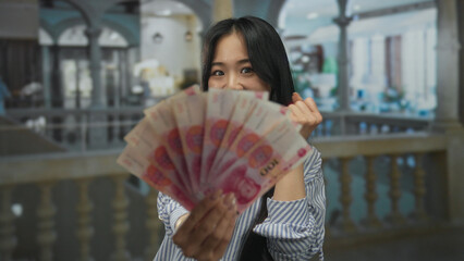 Young woman indoors in a hotel holding chinese yuan banknotes, expressing surprise with a backdrop...