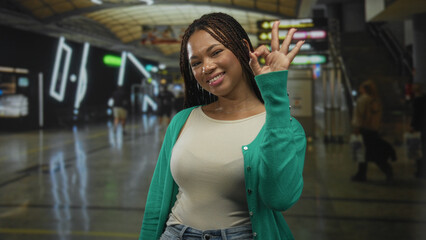 Woman smiling and making ok hand sign in busy airport terminal, showing palm and fingers while wearing green cardigan and jeans; travel confidence.