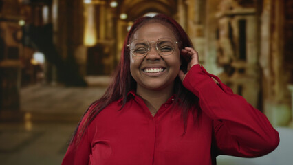 Woman in red shirt and round glasses smiles and touches ear amid stained glass columns in dimly lit church building; warmth joy.