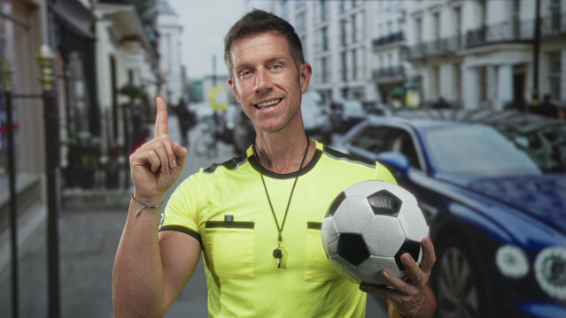 Referee man wearing yellow jersey with whistle holding football points finger up on street; confidence coaching encouragement. - Powered by Adobe