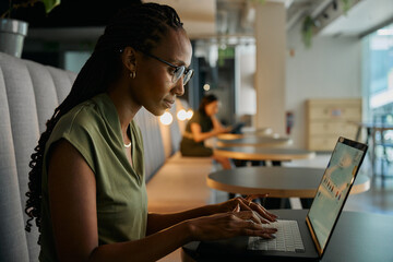 Side view of focused young black businesswoman typing on laptop sitting at desk in office lounge