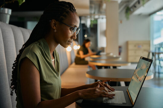 Side view of focused young black businesswoman typing on laptop sitting at desk in office lounge