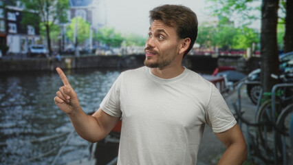 Man pointing finger toward canal on amsterdam street, smiling in a white t shirt with visible...