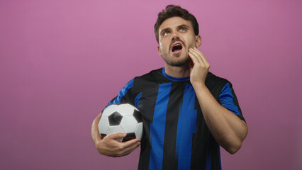 Young hispanic man holding soccer ball and touching cheek with a pained expression, wearing blue...