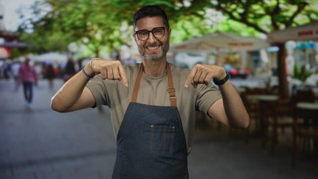 Man points both index fingers to apron while smiling on street terrace restaurant; welcoming small business.