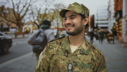 Fototapeta premium Young hispanic man in camouflage military uniform wearing dog tags smiles broadly on crowded city street during daytime; pride duty.