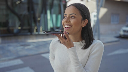 Young woman smiling while using smartphone for voice message on city street during daytime, emphasizing casual and confident communication in an urban environment.