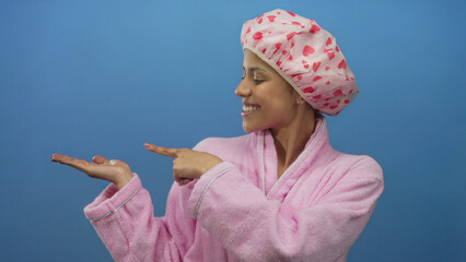 Hispanic woman in pink bathrobe and shower cap smiling and pointing at her palm against a blue...