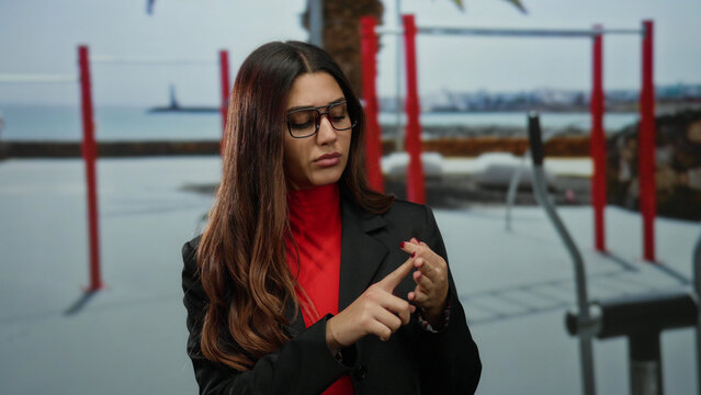 Woman wearing glasses gestures thoughtfully in an outdoor exercise area with red equipment and distant shoreline, dressed in vibrant red and black attire.
