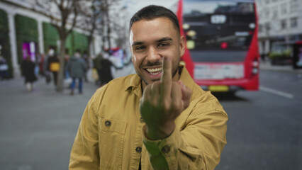 Man raises middle finger gesture on city street with blurred pedestrians and a red bus behind him;...