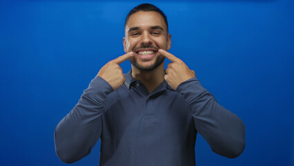 Young man points finger to broad smile with friendly expression in blue studio setting; joy...