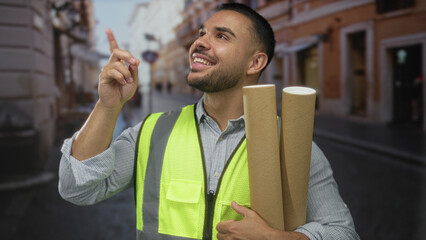 Man holds rolled architectural plans with reflective vest and finger pointing up in urban street;...