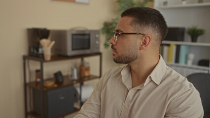 Young hispanic man wearing glasses glances to the side at a coffee station on a shelf in an office...