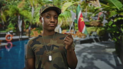 Woman in camouflage uniform and dogtags holding suriname flag at hotel pool outdoors; pride duty.