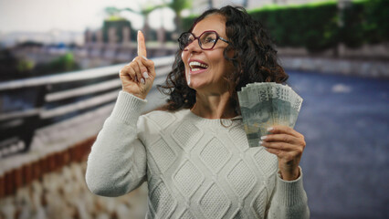Indonesian woman outdoors holding rupiah banknotes smiling with city street background gesturing upwards with finger wearing glasses and white sweater in daylight.
