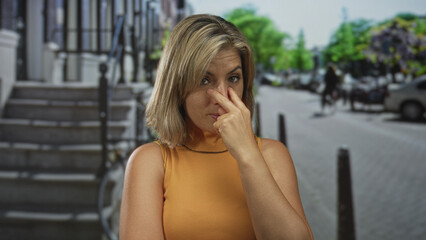 Blonde woman pointing finger forward with poised gesture on a sunlit city street beside stone steps and parked cars; determination perseverance focus resolve.