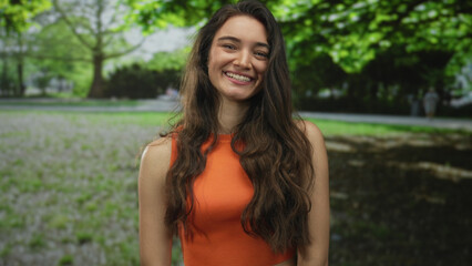 Woman smiling shows bare shoulders and long hair in orange top under green trees in forest bathed...