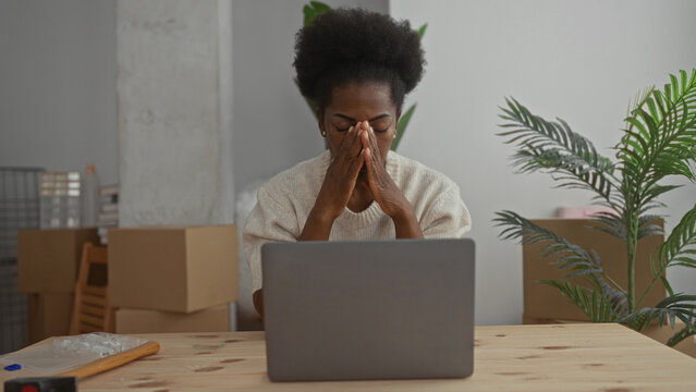 Woman stressed sitting at a desk with laptop in a new home living room surrounded by cardboard boxes and plants implying moving and settling challenges. - Powered by Adobe