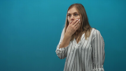 Young blonde woman with finger to lips for silence in blue studio with surprised wide eyed gaze;...