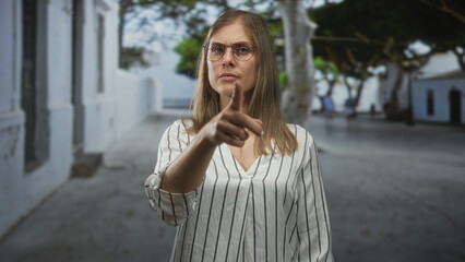Blonde woman wearing glasses pointing finger on street in front of white building wearing striped blouse; confidence.