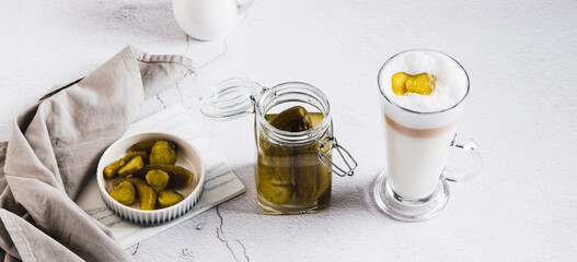 Picklatte with foam in a glass and pickles in a bowl and a jar on the table web banner