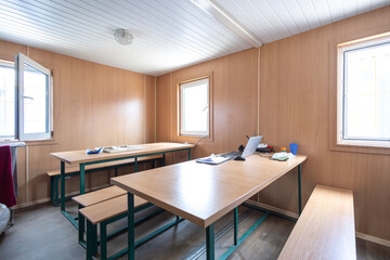 A workers' canteen inside a modular cabin, with long tables and benches for meals. A laptop...