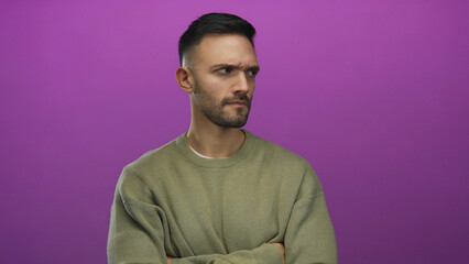 Young hispanic man with serious expression against vibrant pink background in casual sweater arms crossed conveying determination and defiance