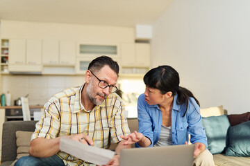 Young couple checking bills and calculating expenses using a laptop at home, debt taxes concept, worried married elderly couple