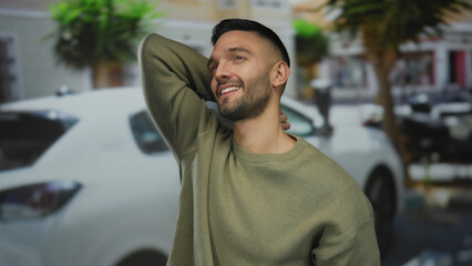 Fototapeta premium Smiling man in sweater stands outdoors on urban street with blurred cars and trees in background evoking a relaxed city vibe.