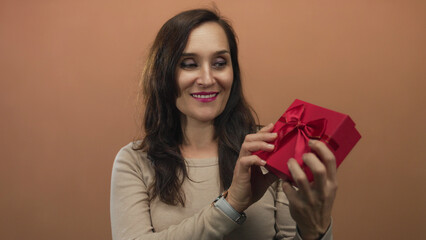 Woman smiling while holding a red gift box against a brown wall showcasing a joyful and festive...