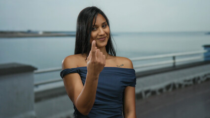 Young hispanic woman gestures come here with her finger on a seaside promenade, with the serene ocean background capturing an inviting and relaxed outdoor ambiance.