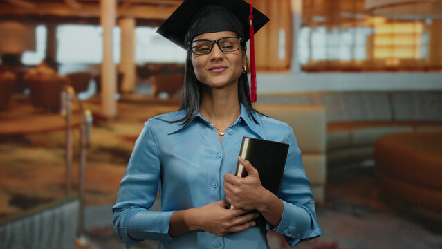 Hispanic woman in graduation cap holding book in modern hotel lobby, smiling confidently at the camera, symbolizing achievement and success in an indoor professional setting.