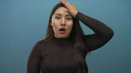 Young hispanic woman with hand on forehead and open mouth against blue studio wall; surprise moment.