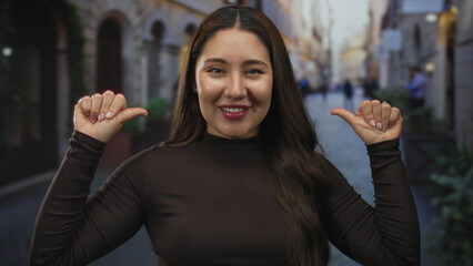 Young hispanic woman pointing thumb to chest on cobblestone street in busy city at dusk; self confidence.