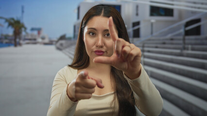 Young hispanic woman holds loser gesture with fingers in front of urban building at seaside...
