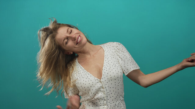 Woman playing air guitar in teal studio with hair flying and bare hands strumming imaginary strings, polka dot dress visible; joy.