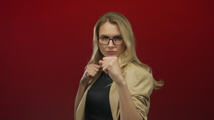 Young blonde woman in glasses stands confidently against red background with fists up, conveying...