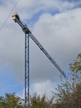blue tower crane with long jib against cloudy sky, tree with green and yellow leaves at bottom right, low angle view of construction site machinery, no people, building industry scene outside