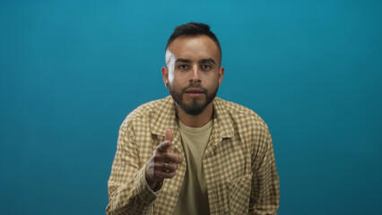 Young man pointing finger to camera with right hand wearing checked shirt in studio against teal backdrop; direct appeal confidence.