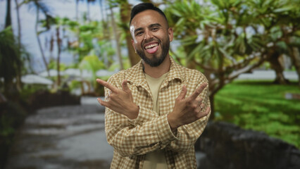 Man smiling with tongue out and showing rock horns with both hands in park wearing checkered beige...